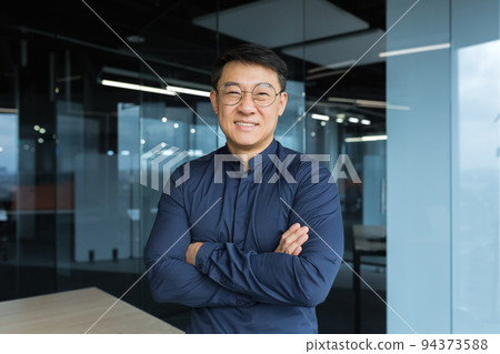 Portrait of happy and successful asian businessman man in glasses smiling and looking at camera, manager business owner with crossed arms near window working inside modern office 94373588