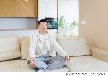 young asian man sits on sofa in lotus position and meditates in don 94375971
