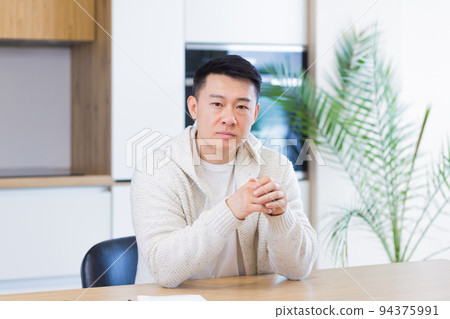 Asian man talking online on video call at home in living room or kitchen. male in casual clothes looking at camera, meeting or conference in distance. Portrait teacher. Lecture training. Webcam view Asian man talking online on video call at home in living room or kitchen. male in casual clothes looking at camera, meeting or conference in distance. Portrait teacher. Lecture training. Webcam view 94375991