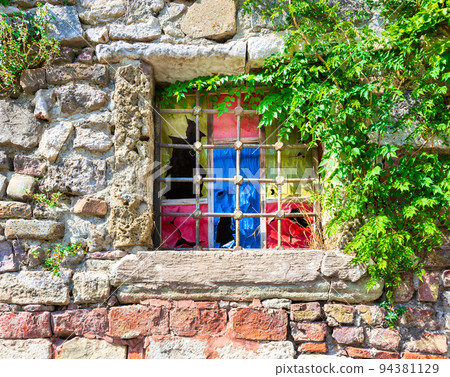 Weathered old stone bricks wall with corroded antique grunge wrought iron window with multi vibrant colored squares Weathered old stone bricks wall with corroded antique grunge wrought iron window with multi vibrant colored squares 94381129