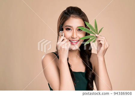 Young ardent woman with healthy skin holding green hemp leaf and cbd oil. Young ardent woman with healthy skin holding green hemp leaf and cbd oil. 94386908