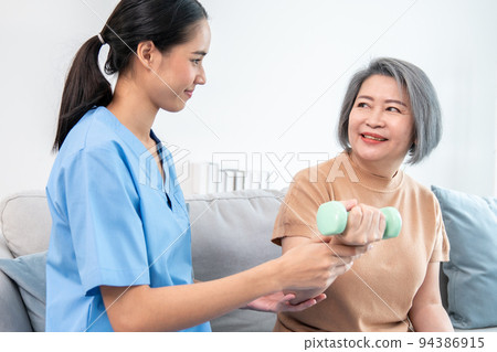 Caregiver helping contented senior woman exercise with dumbbell at home. Caregiver helping contented senior woman exercise with dumbbell at home. 94386915