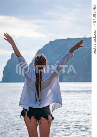 The girl stands on the shore and looks at the sea. Her hands are raised up. She wears a white shirt and her hair is in a braid. 94390460