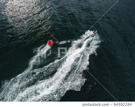 Happy people swim on air mattress behind a high speed water bike. Tourists ride on inflatable watercraft boat. Sea attraction. Water scooter skating people on large Inflatable raft. Aerial copter view 94392284