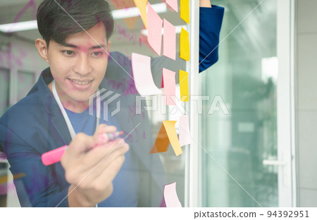 Young Creative businessman holding a marker and writing plan and share idea on glass wall with sticky note, Brainstorming and discussing and formulating, business strategies in tech startup office. 94392951