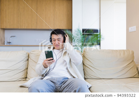 young cheerful asian guy listening to music in headphones sitting on sofa at home. A man alone on the couch feels good lifestyle. Happy male with the phone listens to his favorite radio station online young cheerful asian guy listening to music in headphones sitting on sofa at home. A man alone on the couch feels good lifestyle. Happy male with the phone listens to his favorite radio station online 94393322