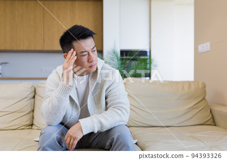 young asian man sitting on sofa at home pensive, worried about problems and depression. male has an experience relationships at work or family, problems with loan repayment debt Seriously conceived 94393326