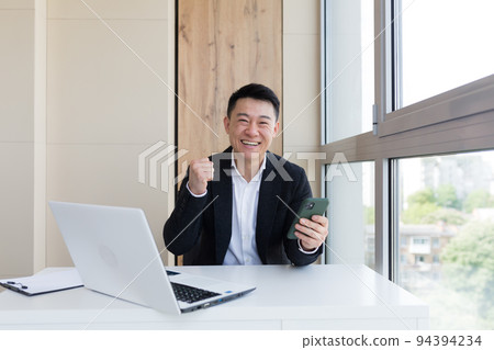 Happy young Asian business man in office, looking at camera, with emotions of winner or victory, financial promotion sports betting. The man exclaims happily while playing the game. Excited with joy 94394234