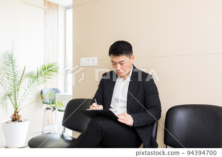 Asian businessman man, in the waiting room sitting on a chair near the reception of the office center, and fills out a test form to receive his application Asian businessman man, in the waiting room sitting on a chair near the reception of the office center, and fills out a test form to receive his application 94394700