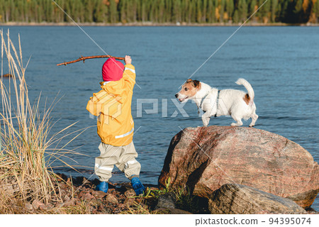 Little girl wearing bright and colourful waterproof clothing and rubber boots throwing wooden stick to dog. Family playing at Fall beach of nothern sea. Polar circle, White Sea, Karelia. Little girl wearing bright and colourful waterproof clothing and rubber boots throwing wooden stick to dog. Family playing at Fall beach of nothern sea. Polar circle, White Sea, Karelia. 94395074