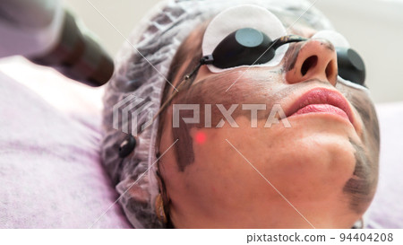 A woman in protective glasses with a smeared black mask on her face is undergoing a procedure in a beauty salon A woman in protective glasses with a smeared black mask on her face is undergoing a procedure in a beauty salon 94404208
