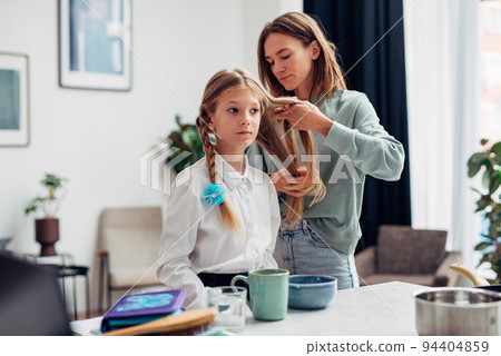 Mother braids her daughter's braids before school at breakfast 94404859