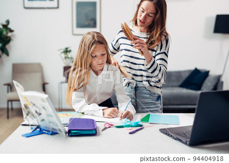 Mother brushes a girls hair while doing her homework. Mother brushes a girls hair while doing her homework. 94404918
