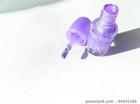 An open bottle with lilac nail polish and a brush next to it on a white surface with a shadow, the concept of fashionable beauty 94405388