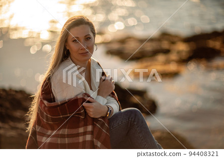 Attractive blonde Caucasian woman enjoying time on the beach at sunset, sitting in a blanket and looking to the side, with the sunset sky and sea in the background. Beach vacation. Attractive blonde Caucasian woman enjoying time on the beach at sunset, sitting in a blanket and looking to the side, with the sunset sky and sea in the background. Beach vacation. 94408321