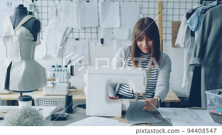 Young attractive dressmaker is working on sewing machine and looking at women's garment sketches in her light studio at table. Modern equipment and fashionable garments in background. 94408547