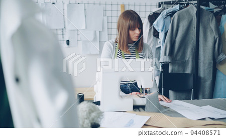 Self-employed female clothing designer is working with sewing machine, stitching textile and looking at sketches lying on studio table. Everyday work at tailor's shop concept. Self-employed female clothing designer is working with sewing machine, stitching textile and looking at sketches lying on studio table. Everyday work at tailor's shop concept. 94408571