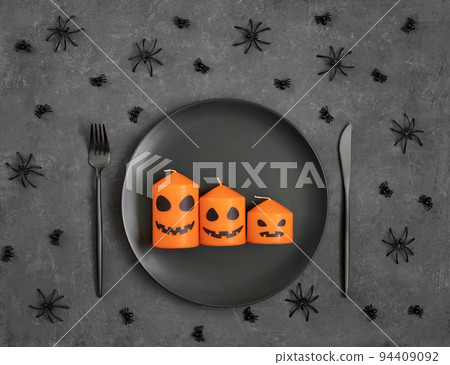 Halloween table setting with black dishes and jack lantern candles in a plate, spiders on a dark gray background. Festive serving. Top view, flat lay. 94409092