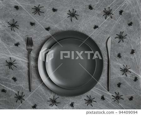 Halloween table setting with black crockery and spiders in the web. Empty black plate on a gray background. Halloween festive serving. Copy space, top view, flat lay. 94409094