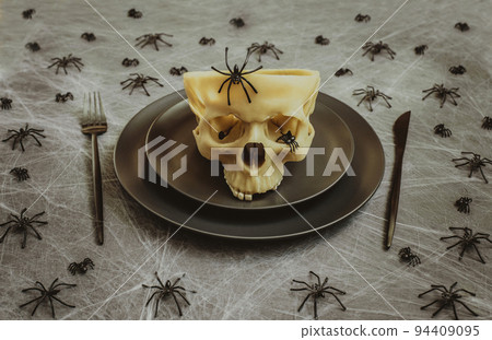 Halloween table setting with black cutlery, skull and spiders in a web on a gray background. Creative festive serving. Halloween table setting with black cutlery, skull and spiders in a web on a gray background. Creative festive serving. 94409095