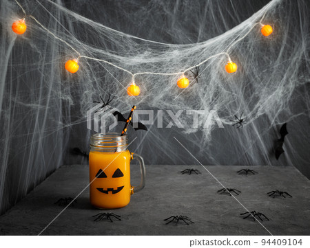 Halloween pumpkin juice in a glass jar with a scary face, spiders and cobwebs on a dark gray background. Funny decorations for a children's party. Copy space. Halloween pumpkin juice in a glass jar with a scary face, spiders and cobwebs on a dark gray background. Funny decorations for a children's party. Copy space. 94409104