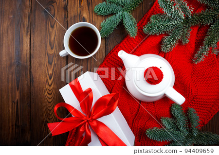 On a wooden background there is a red scarf and there is a teapot and a cup of tea 94409659