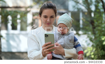Mom with her son in her arms stands against the backdrop of a multi-storey building in the yard and shows him a cartoon or a funny children's video Mom with her son in her arms stands against the backdrop of a multi-storey building in the yard and shows him a cartoon or a funny children's video 94410330