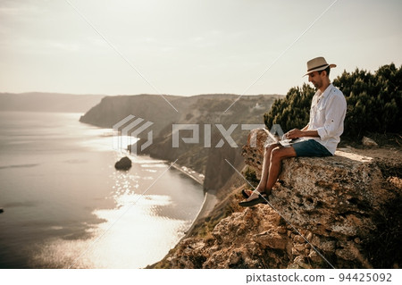 Digital nomad, man in the hat, a businessman with a laptop sits on the rocks by the sea during sunset, makes a business transaction online from a distance. Remote work on vacation. 94425092