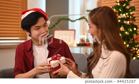 Young man giving surprise gift to his girlfriend during celebrating New Year at home. Family, holidays and people concept Young man giving surprise gift to his girlfriend during celebrating New Year at home. Family, holidays and people concept 94425287