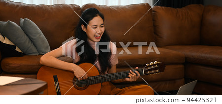 Happy young woman learning play guitar, watching online lessons on laptop. Hobbies and learning concept Happy young woman learning play guitar, watching online lessons on laptop. Hobbies and learning concept 94425321