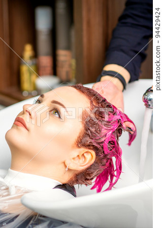 Washing dyed female hair. A young caucasian woman having her hair washed in a beauty salon. Professional hairdresser washes pink color paint off of a customer's hair Washing dyed female hair. A young caucasian woman having her hair washed in a beauty salon. Professional hairdresser washes pink color paint off of a customer's hair 94429424