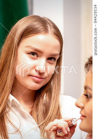 Makeup in the process. The makeup artist applies pink gloss lipstick on the lips of the beautiful face of the young caucasian woman in a beauty salon Makeup in the process. The makeup artist applies pink gloss lipstick on the lips of the beautiful face of the young caucasian woman in a beauty salon 94429534