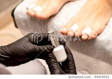 White nail polish in the hands of a manicurist while painting nails on a female feet, closeup 94429581