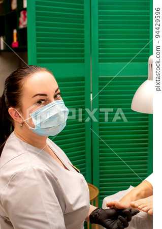 Young female podiatrist with a protective mask doing chiropody and looking at the camera in her podiatry clinic Young female podiatrist with a protective mask doing chiropody and looking at the camera in her podiatry clinic 94429596