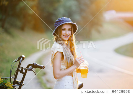 A happy girl in white shorts and a yellow blouse holds a glass of juice with a straw on the background of the road near the forest A happy girl in white shorts and a yellow blouse holds a glass of juice with a straw on the background of the road near the forest 94429998