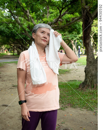 Happy senior Asian woman smiling standing on grass with sports towel around neck enjoying nature fresh air in park. 94430592