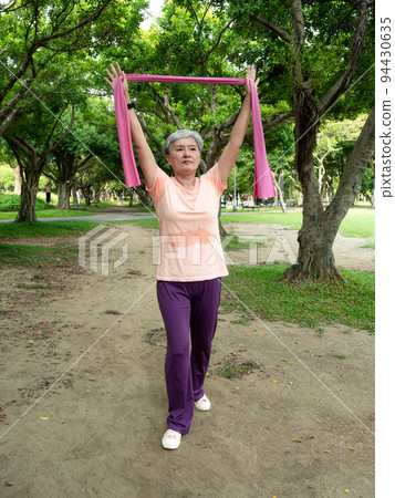 Happy senior Asian woman smiling and standing on grass using elastic band for exercise and enjoying nature fresh air in park. 94430635