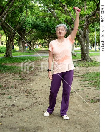 快樂的樂齡亞洲婦女微笑著站在草地上使用啞鈴運動,並且享受公園裡的大自然新鮮空氣. 94430701