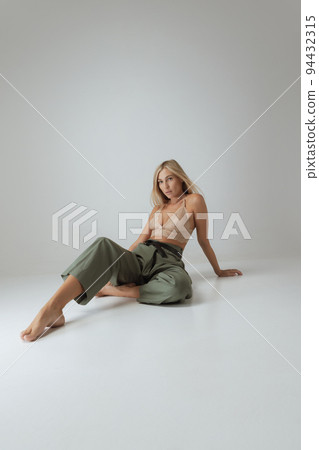 Beautiful woman with long blond hair and natural makeup wearing lacy top and casual trousers sitting on floor isolated on grey background. Beautiful woman with long blond hair and natural makeup wearing lacy top and casual trousers sitting on floor isolated on grey background. 94432315