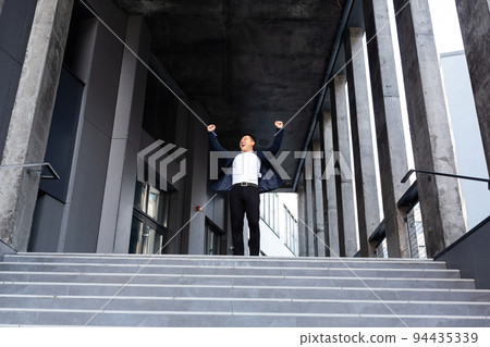 Happy and cheerful asian businessman holding hands up rejoicing in victory and success near office center front view Happy and cheerful asian businessman holding hands up rejoicing in victory and success near office center front view 94435339