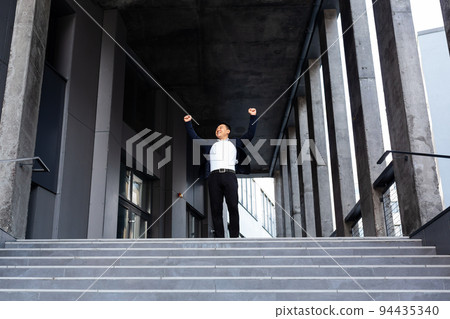 Happy and cheerful asian businessman holding hands up rejoicing in victory and success near office center front view Happy and cheerful asian businessman holding hands up rejoicing in victory and success near office center front view 94435340