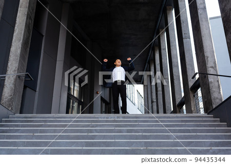 Happy and cheerful asian businessman holding hands up rejoicing in victory and success near office center front view Happy and cheerful asian businessman holding hands up rejoicing in victory and success near office center front view 94435344