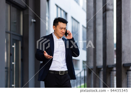 angry and serious Successful Asian businessman explains information to employees using phone, speaks near office outside angry and serious Successful Asian businessman explains information to employees using phone, speaks near office outside 94435345