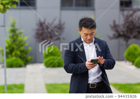 Asian businessman boss looks at mobile phone reads news from phone and writes in messenger outside office Asian businessman boss looks at mobile phone reads news from phone and writes in messenger outside office 94435926