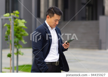 Asian businessman boss looks at mobile phone reads news from phone and writes in messenger outside office Asian businessman boss looks at mobile phone reads news from phone and writes in messenger outside office 94435927