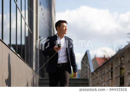 Asian businessman boss looks at mobile phone reads news from phone and writes in messenger outside office Asian businessman boss looks at mobile phone reads news from phone and writes in messenger outside office 94435928