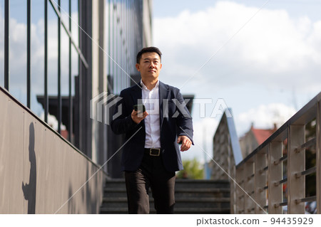 Asian businessman boss looks at mobile phone reads news from phone and writes in messenger outside office Asian businessman boss looks at mobile phone reads news from phone and writes in messenger outside office 94435929