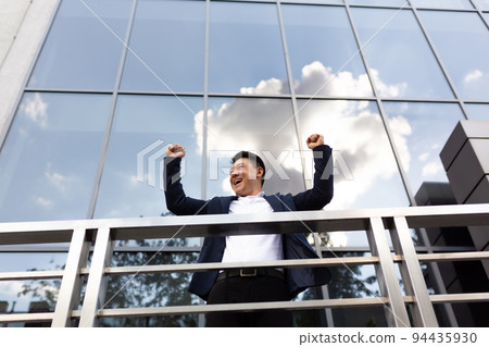 Asian businessman boss stands near his office center on the balcony in a dark color business suit holds his hands up celebrates victory Asian businessman boss stands near his office center on the balcony in a dark color business suit holds his hands up celebrates victory 94435930