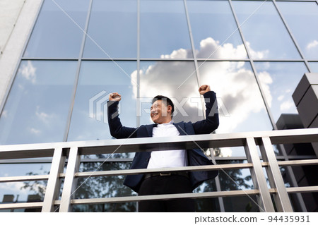 Asian businessman boss stands near his office center on the balcony in a dark color business suit holds his hands up celebrates victory Asian businessman boss stands near his office center on the balcony in a dark color business suit holds his hands up celebrates victory 94435931