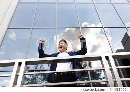 Asian businessman boss stands near his office center on the balcony in a dark color business suit holds his hands up celebrates victory Asian businessman boss stands near his office center on the balcony in a dark color business suit holds his hands up celebrates victory 94435932